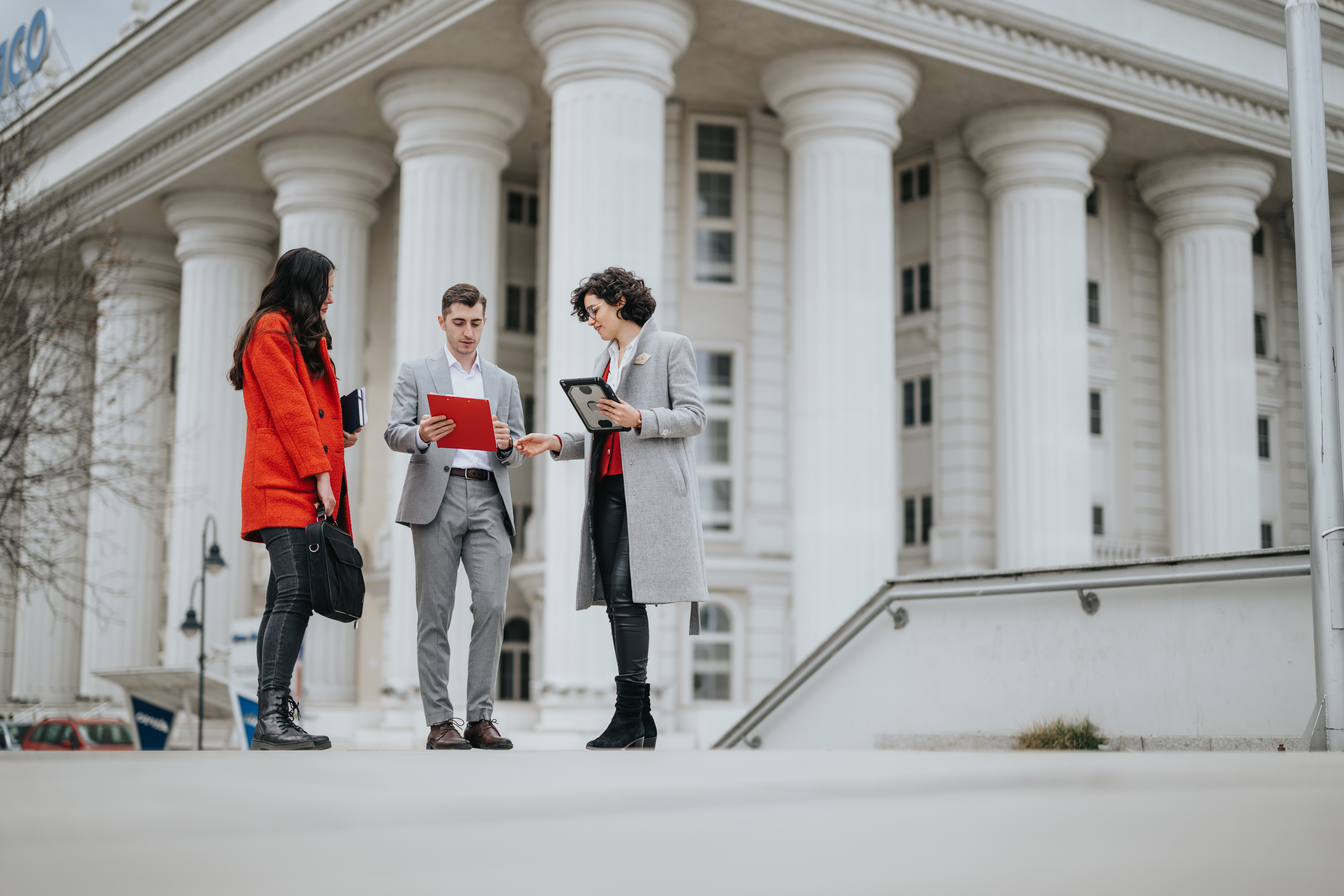 Three people standing in front of a government building looking at a document the person in the middle is holding.