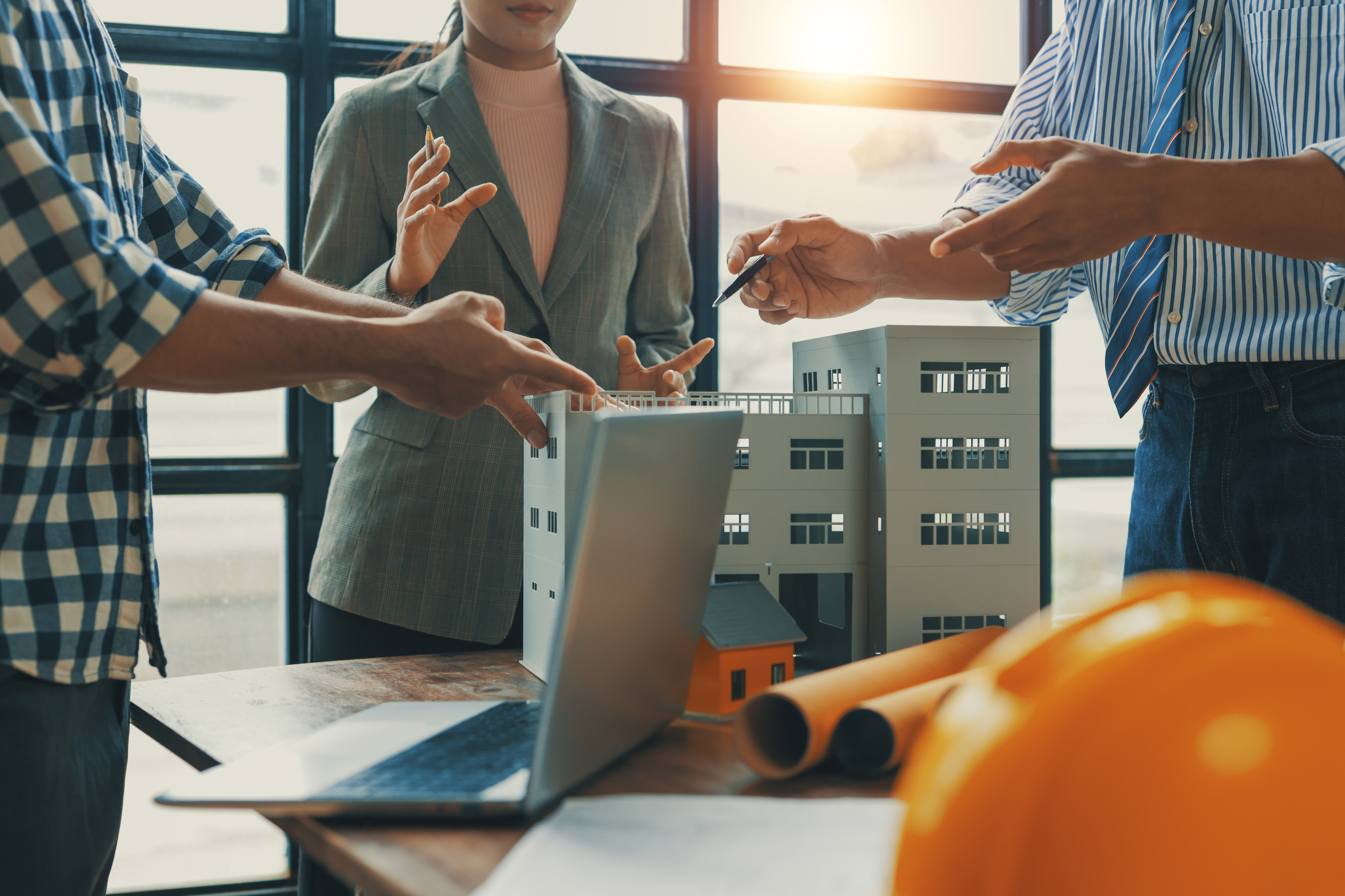 3 people discussing a commercial building model over a table with laptop and blueprints in a sunny office