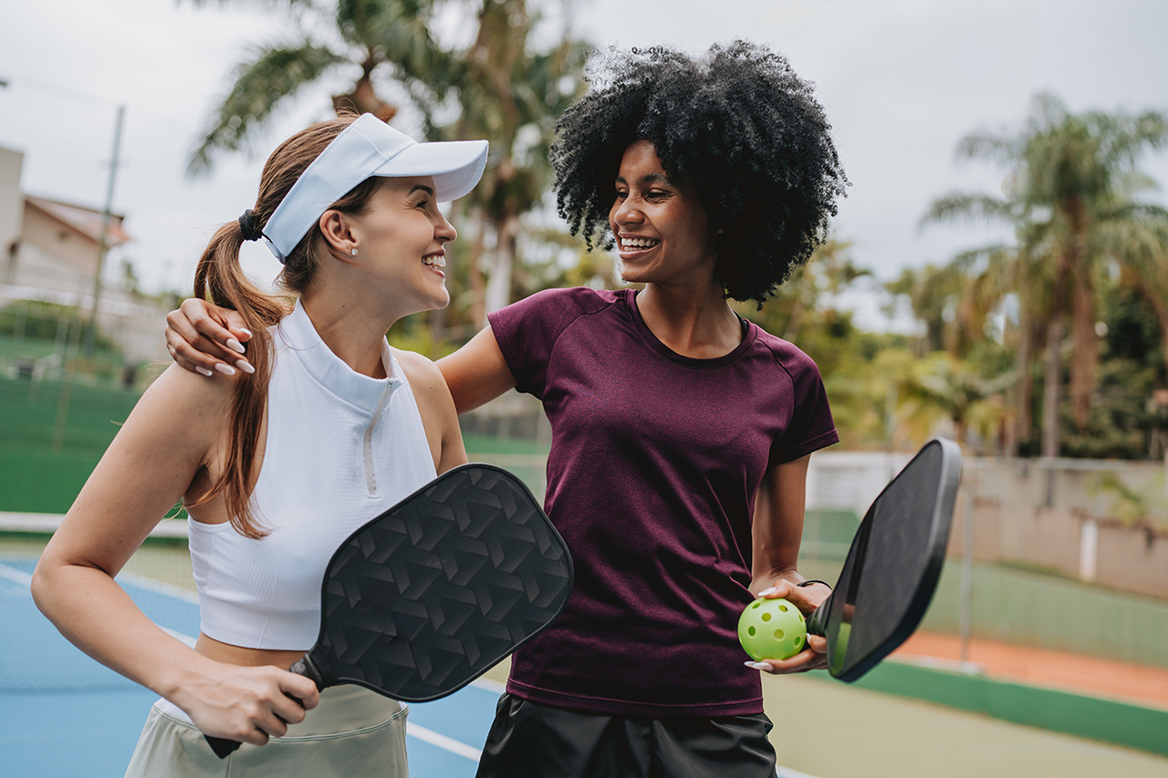Two women playing pickleball on an outdoor court, smiling at each other