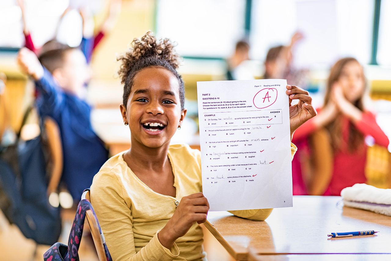 Child holding up a school paper with a smile, surrounded by classmates