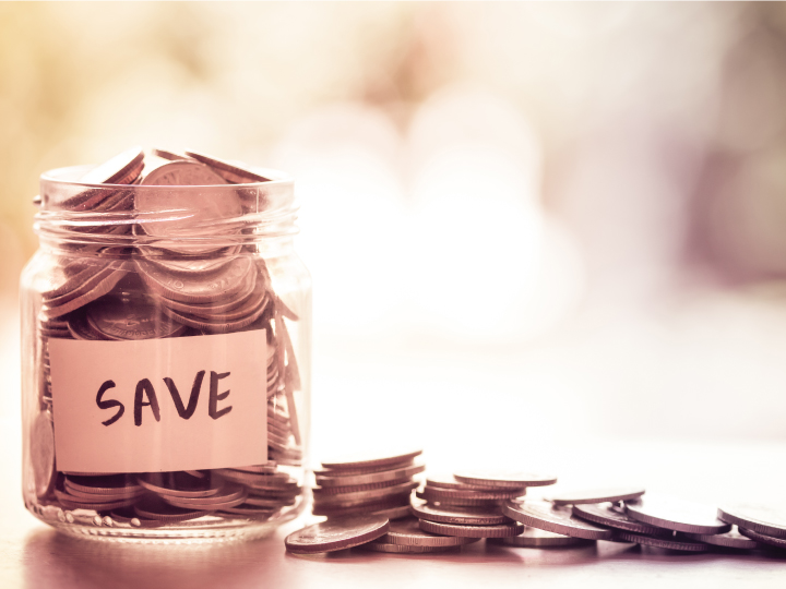 glass jar on table filled with coins with additional coins on the table and blurry background 